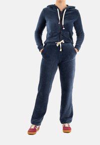 Chándal de pana azul marino con sudadera con cremallera y cinturilla con cordón. Presenta una textura acanalada y bolsillos frontales. Combinado con zapatillas rojas.