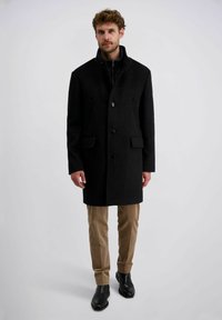 Langer schwarzer Wollmantel mit hohem Kragen, zwei vorderen Taschen und Knopfverschluss, kombiniert mit beigen Hosen und schwarzem Schuhwerk.