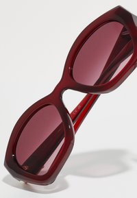 Κόκκινα υπερμεγέθη γυαλιά ηλίου με παχύ, γυαλιστερό σκελετό και σκούρους κόκκινους φακούς. Διαθέτουν γωνιώδεις σχήματα και λεία υφή.