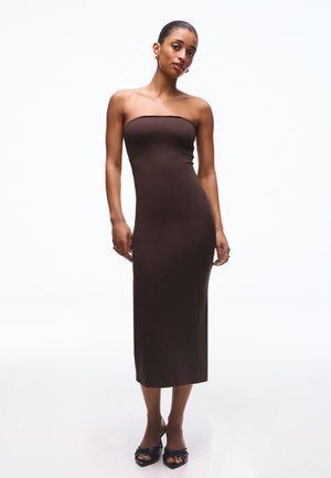 Femeie îmbrăcată într-o rochie midi maro, fără bretele, mulată, purtând pantofi negri cu toc și vârf decupat, în fața unui fundal alb simplu.