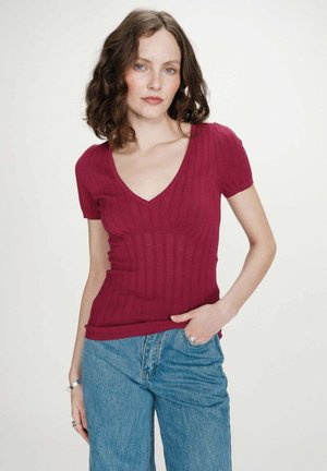 Femme aux cheveux bouclés bruns portant un haut en maille rouge foncé moulant à manches courtes et un jean bleu, debout devant un fond blanc uni.