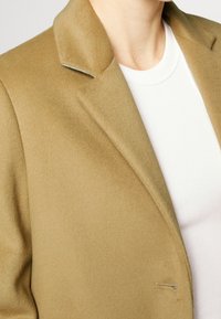 Blazer in misto lana beige con colletto a punta e dettaglio a un bottone. Indossato sopra una maglietta bianca a costine. Texture morbida visibile.