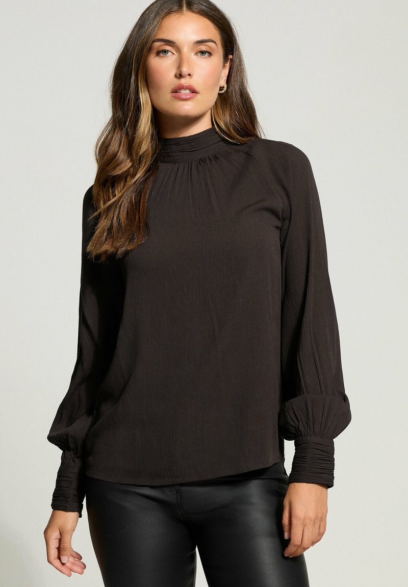 Mustassa pitkähihaisessa paidassa on pleidattu korkeakaulus ja kootut hihansuut. Pehmeä, teksturoitu kangas; rento malli, jossa on hieman pyöristetty helma.