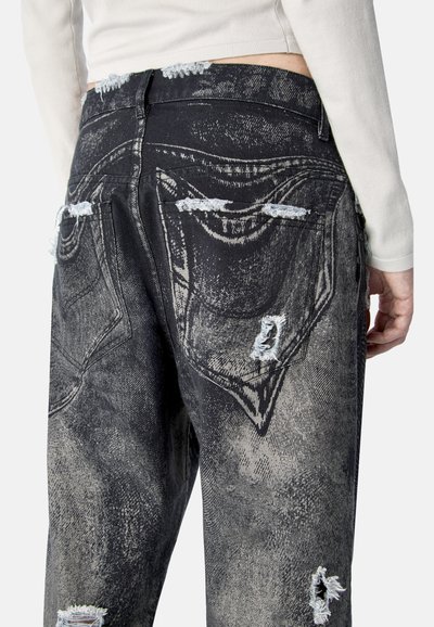 Vista trasera de una persona vistiendo jeans negros con efecto de lavado ácido y rotos desgastados, combinados con un top de manga larga color crema, con la mano relajada al costado.