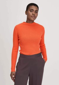 Orange gestreiftes Langarmoberteil mit hohem Kragen, kombiniert mit dunklen, locker sitzenden Hosen. Glatte Textur und figurschmeichelndes Design.