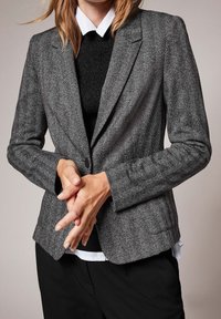 Blazer gris à motif chevrons avec une coupe ajustée, des revers crantés et une fermeture à un bouton, associé à un pull noir et une chemise blanche en dessous.