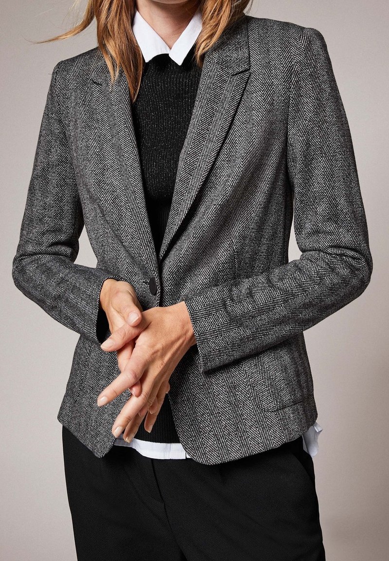 Blazer gris à motif chevrons avec une coupe ajustée, des revers crantés et une fermeture à un bouton, associé à un pull noir et une chemise blanche en dessous.