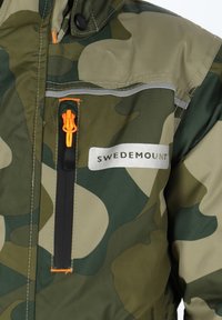 Camo jack in groen en beige met een oranje ritssluiting. Bevat een gestikt label met de tekst "SWEDEMOUNT" en reflecterende accenten.
