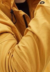 Žlutá lehká bunda se zipovým uzávěrem, nastavitelným stahovacím šňůrkem a texturovanou látkou. Na límci má černý knoflík jako ozdobu.