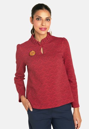 Rote Bluse mit Muster, Rüschenausschnitt, Schlüssellochausschnitt und floralem Broschenakzent. Lange Ärmel und glatter Stoff.