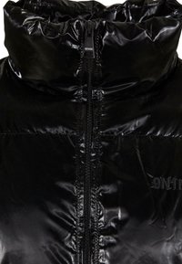 Doudoune noire avec un col haut, dotée d'une fermeture éclair à l'avant et d'un tissu brillant et texturé. Le logo de la marque est brodé sur le côté.