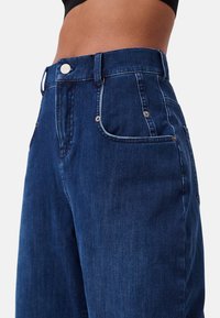 I jeans in denim blu scuro presentano una vita alta, tasche anteriori e dettagli in metallo. Il tessuto ha una superficie liscia e dettagli di cucitura minimi.