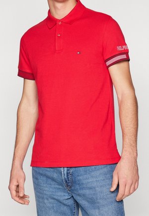 Mand iført en rød kortærmet polo med stribede manchetter og lille logo på brystet, kombineret med blå jeans.