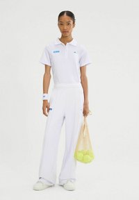 Camisa de polo blanca de manga corta, con logotipos azules. Combinada con pantalones anchos blancos y una bolsa de malla con pelotas de tenis verdes.