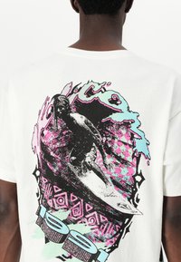White short-sleeve t-shirt featuring a colorful graphic of a kneeboarder on waves, with geometric patterns and accents in pink and aqua.