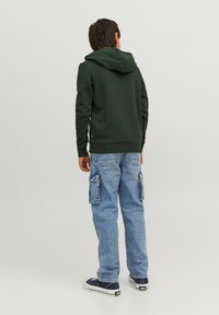 Felpa verde scuro con tasca posteriore, abbinata a jeans cargo azzurri chiari con tasche laterali e una vestibilità casual. Sneakers di colore blu navy.