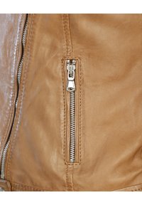 Beige Lederjacke mit seitlicher Reißverschlusstasche und silberner Hardware, glatter Textur und sichtbaren Nähten.