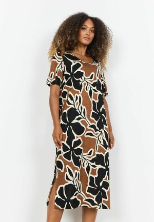 Ženska s kodrastimi lasmi, oblečena v rjavo midi obleko z velikimi črno-belimi cvetličnimi vzorci, stoji pred enakomerno svetlo ozadje.