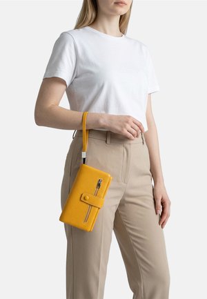 Donna che indossa una maglietta bianca e pantaloni beige tiene una pochette gialla testurizzata con chiusura a cerniera e bottone a pressione.