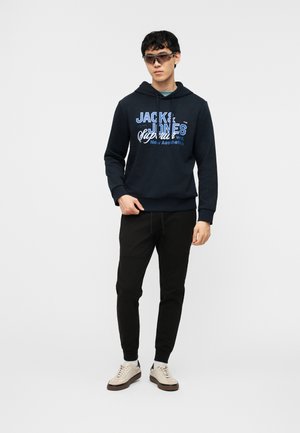 Ung mann iført svarte solbriller, navy hettegenser med teksten "Jack & Jones", svarte joggebukser og beige joggesko, som står foran en hvit bakgrunn.
