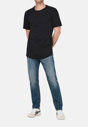 Mies, joka pukeutunut yksinkertaiseen mustaan lyhythihaiseen t-paitaan, sinisiin farkkuihin ja valkoisiin lenkkareihin, seisoo valkoista taustaa vasten kädet selän takana.