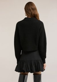 Musta lyhyt neulepaita, jossa on resoripinta, leveät hihat ja viittakaulus. Yhdistetty leveälahkeiseen mustaan hameeseen ja kiiltäviin polven yli ulottuviin saappaisiin.