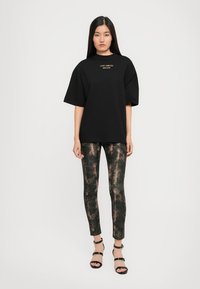 Maglietta nera oversize con testo dorato, abbinata a leggings verde scuro con motivo a pelle di serpente. Sandali neri con lacci completano l'outfit.
