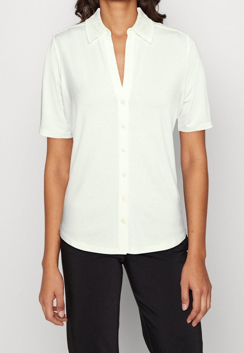 Femme portant une chemise blanche à manches courtes avec col et des boutons, associée à un pantalon noir, se tenant devant un fond uni.