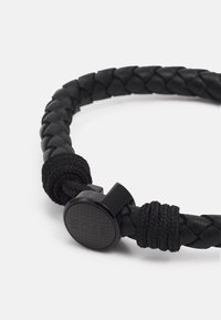 Pulsera de cuero trenzado negro con un cierre de metal texturizado. Presenta un detalle trenzado y un cierre circular liso.