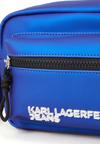 Blå nylonveske med en glatt tekstur, med en svart glidelås, sølvfarget maskinvare og hvit preget tekst som sier "KARL LAGERFELD JEANS."