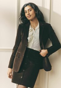 Barna kordbársony blézer és hozzáillő szoknya, alattuk fehér fodros blúz. A blézer elején zsebek és gombok; az egész anyag sima tapintású.