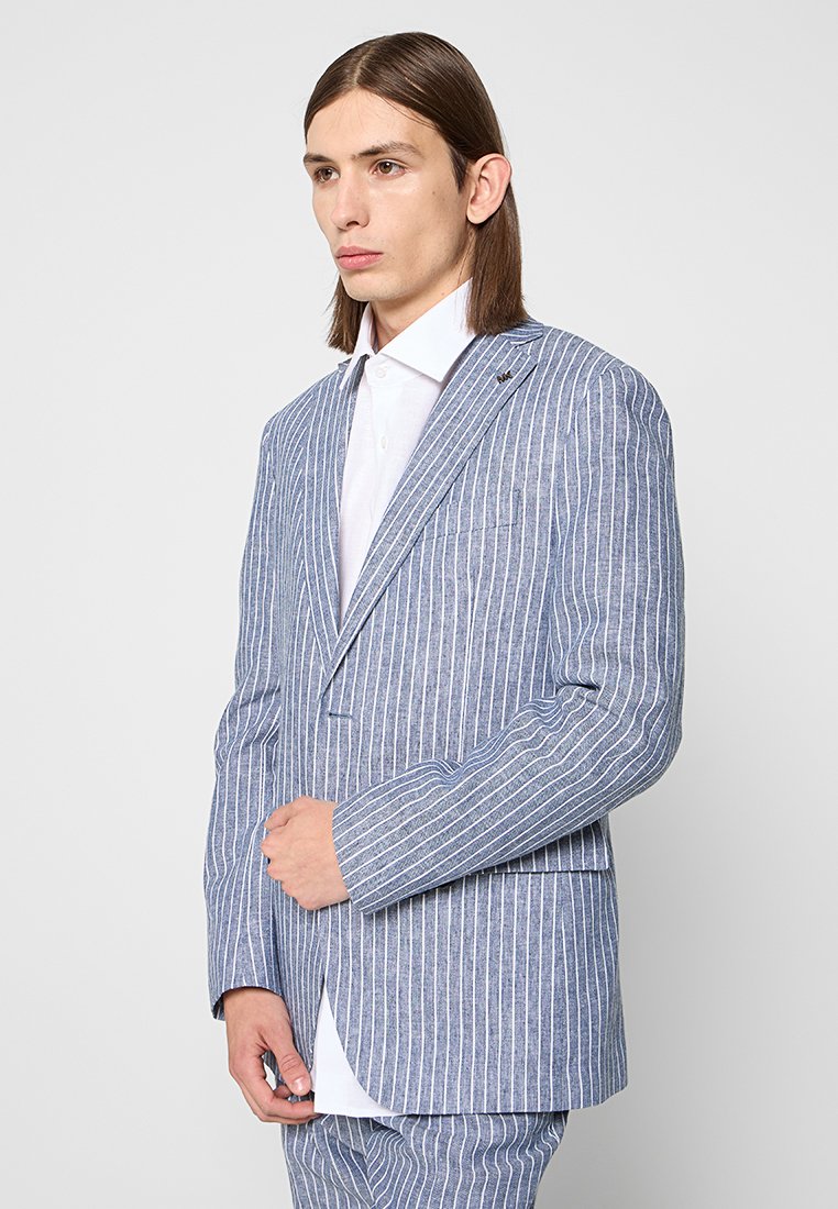 Młody mężczyzna z włosami do ramion, ubrany w niebieski i biały garnitur w pionowe paski oraz białą koszulę z guzikami.