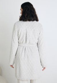 Rochie căptușită de culoare gri deschis, cu mâneci lungi și talie legată, având un model texturat în formă de diamant și material moale, pufos.