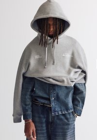 Grå bomullshuvtröja med huva, med vit text framtill, lagd över en blå denimskjorta och matchande jeans.