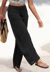 Schwarze, weite Hosen aus leichtem Stoff mit elastischem Bund. Kombiniert mit schwarzen Sandalen mit goldenen Akzenten und einer geflochtenen Tragetasche.