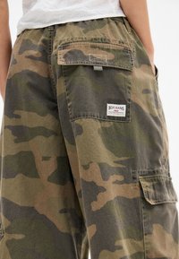 Calças cargo camufladas em tons de verde e castanho, com múltiplos bolsos e um rótulo da BDG Jeans. Feitas de um tecido resistente, com um corte descontraído.