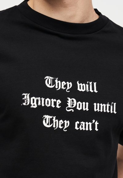 Μαύρο T-shirt με λευκό γοτθικό κείμενο που γράφει «Θα σε αγνοούν μέχρι να μην μπορούν» φορεμένο από άτομο που φαίνεται από τον λαιμό έως τη μέση του κορμού.