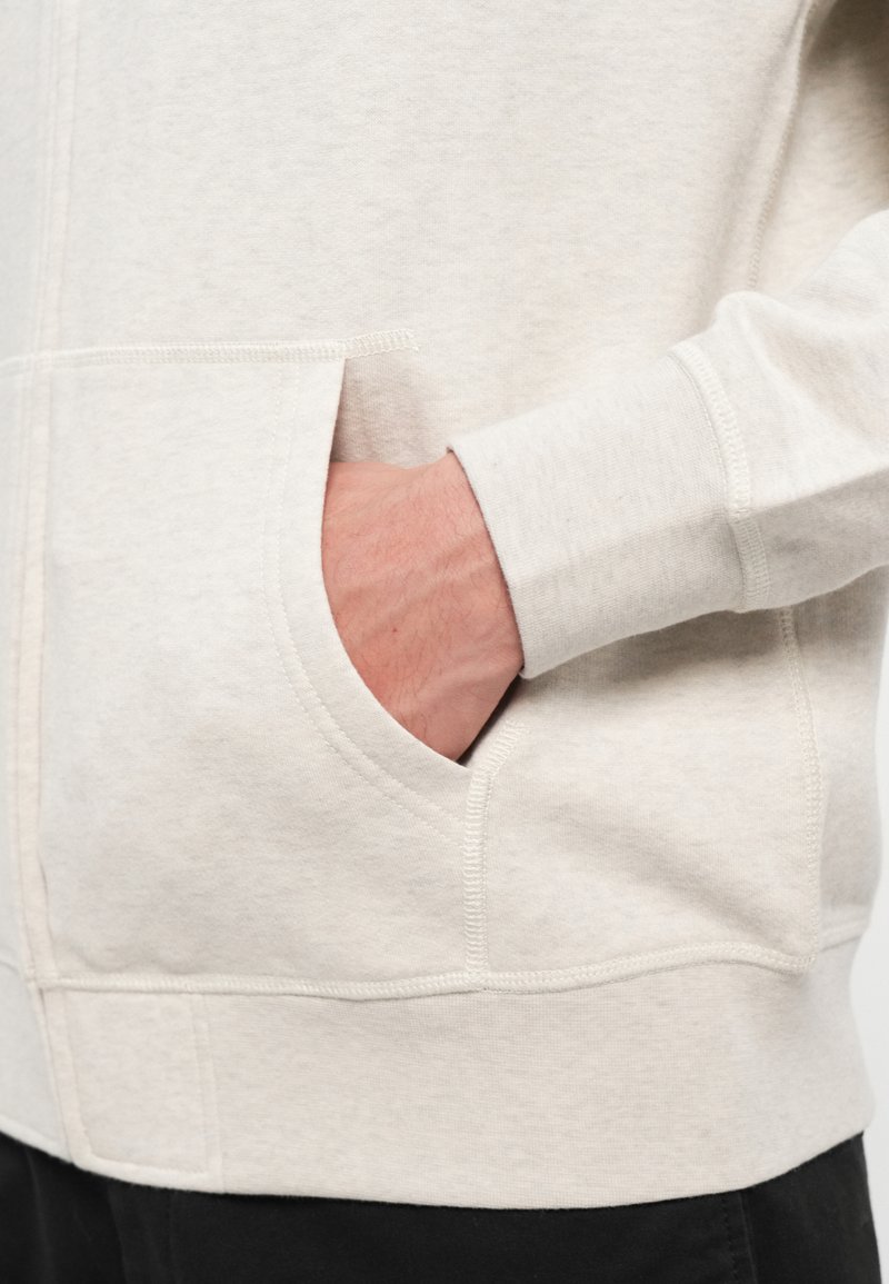 Hand teilweise in der vorderen Tasche eines hellbeigen Hoodies, der über schwarzen Hosen getragen wird, wobei die Stoffstruktur und die Nähdetails sichtbar sind.