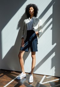 Blazer bianco sopra una camicia bianca, abbinato a pantaloni corti scuri. Il modello indossa sneakers bianche. Luce e ombra creano motivi distintivi sulla parete.