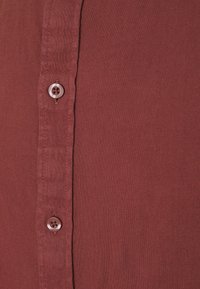 Bordeaux knappet skjorte med struktureret stof, en række knapper og en synlig søm langs siden. Stoffet ser ud til at være åndbart.