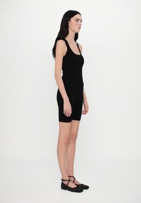 Combinaison sans manches noire au design ajusté, s'étendant jusqu'à mi-cuisse, portée avec des ballerines noires cloutées avec des brides entrecroisées.
