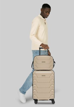 Man in beige lange mouwen shirt en spijkerbroek die een beige SMARTBOX-koffer met uitschuifbare handgreep vasthoudt, staand tegen een effen grijze achtergrond.