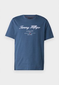 Kék pamut póló, kerek nyakkal és rövid ujjakkal, fehér nyomtatott szöveggel: "Tommy Hilfiger 1985-BEN ALAPÍTVA NEW YORK."