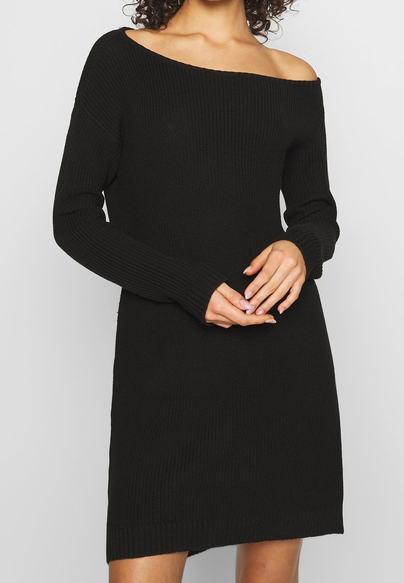 Femme portant une robe noire en maille à épaules dénudées avec manches longues, mains doucement jointes à la taille, sur fond gris clair.