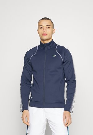 Homme portant une veste zippée bleue marine avec des rayures blanches sur les manches et un pantalon blanc, se tenant devant un fond uni clair.