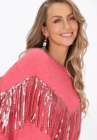 Różowy sweter z dzianiny z frędzlami i metalicznymi cekinami wzdłuż dołu. Elegancka faktura i żywy kolor podkreślają styl.