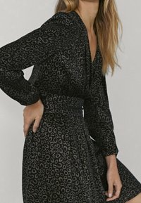Robe noire à manches longues, avec un motif léopard dans des nuances de noir et d'or, taille cintrée et texture de tissu douce.