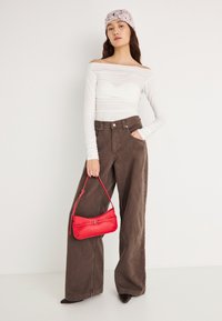 Witte off-shoulder top met lange mouwen, bruine broek met wijde pijpen en een rode leren handtas met korte hengsel, gedragen met zwarte pumps met een spitse neus.