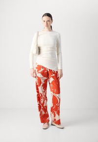 White long-sleeve top, fitted. Red floral-patterned wide-leg pants. Light-colored flat shoes. Model holds a light bag. Simple background.