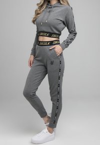 Grijze cropped hoodie en joggers van zachte stof, met zwarte en gouden SIK SILK branding, zijstrepen en witte sneakers.